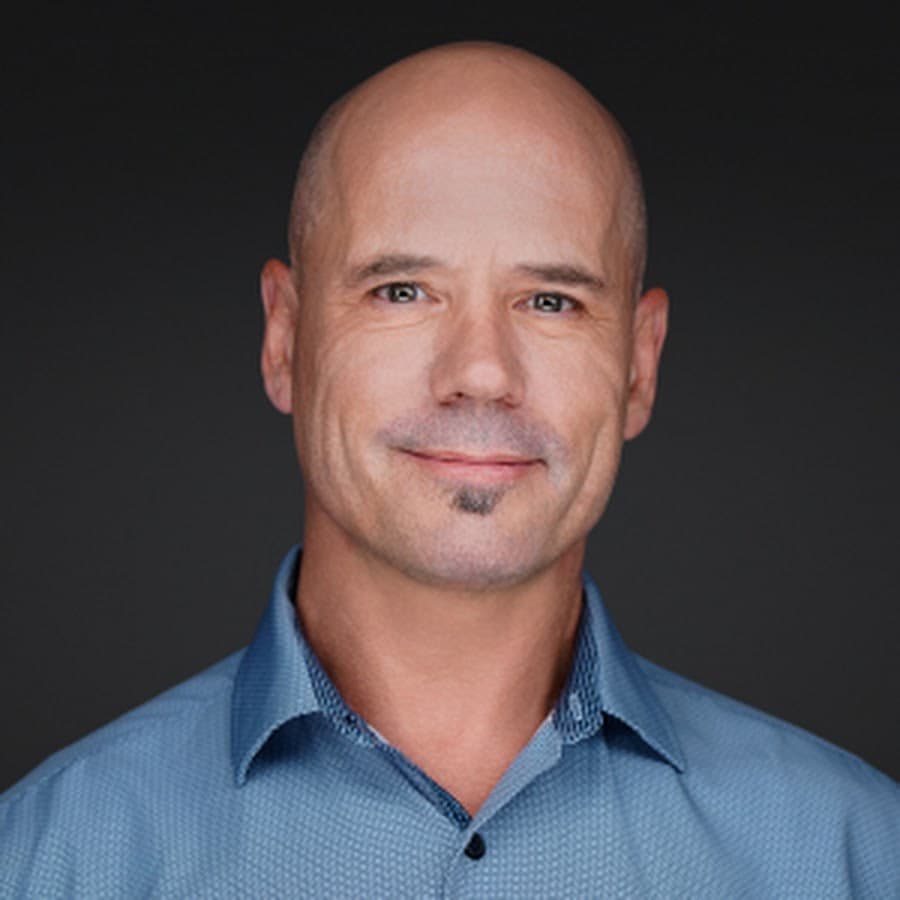 Smiling bald man with a soul patch wearing a blue shirt against a dark background.